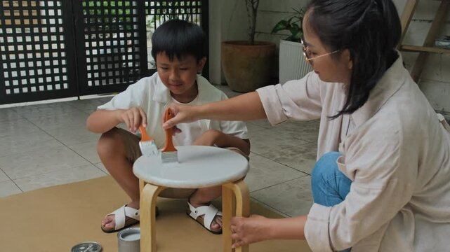 Medium long shot of Asian mom and her teenage son using brush while painting old stool in backyard