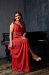 A woman in a red dress sits on a stool in front of a piano. She is wearing a necklace and a ring. Scene is elegant and sophisticated