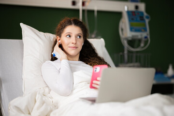 Using gadgets in bed, the girl in the hospital holds a phone and works on a computer.