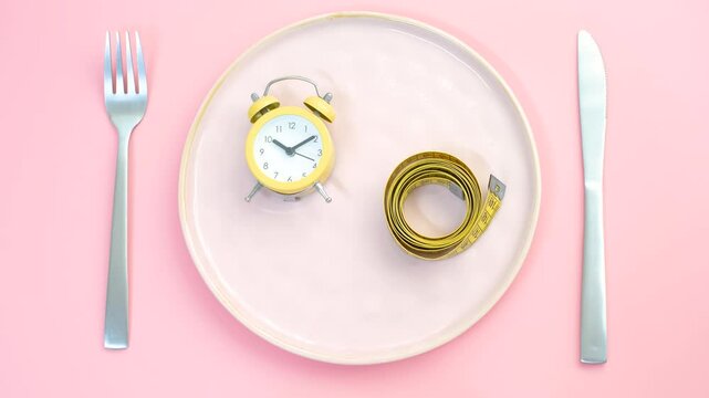 Empty plate with alarm clock and measuring tape on pink background, intermittent fasting concept.