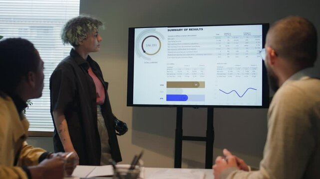 Girl with bionic prosthetic arm showing summary of results of business campaign pointing at digital screen, two multi-ethnic male coworkers watching