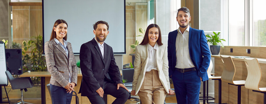 Team of young successful business professionals at work meeting. Four happy people in office. Banner with group portrait of cheerful men and women in classic suits looking at camera and smiling