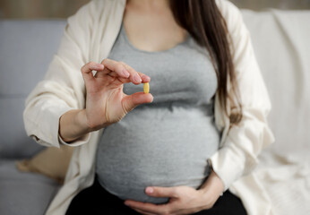 Close up of young pregnant woman holding pill in hand, taking vitamins D, E, A, calcium, dietary supplements at home. Pregnancy, prenatal health and wellness, healthy nutrition, hydration