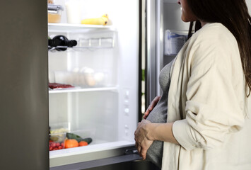 Thoughtful hungry pregnant woman standing in front of open fridge, refrigerator in middle of night at home in kitchen, closeup. Healthy maternity eating, nutrition, pregnancy diet. Looking for food