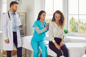 Woman nurse with stethoscope examining female patient lungs, breathing and heartbeat at the hospital in exam room with male doctor standing next to them in clinic. Medical checkup and health care.