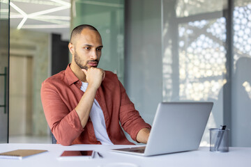 Thoughtful man with laptop in modern office. Professional analyzing work strategy. Wearing casual...