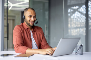 Smiling professional wearing headset works on laptop in modern office setting. Engaged in online meeting using phone technology. Notebook and pen on table indicate readiness and focus.