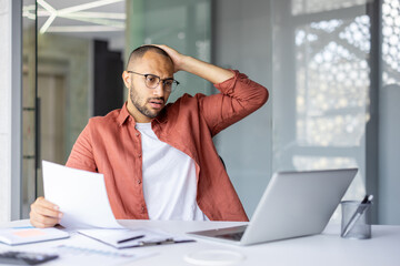 Worried man in office holding document while looking at laptop with stressed expression. Professional setting suggests business concern, stress, and decision-making scenario.