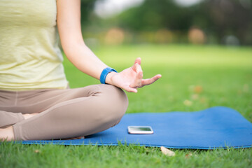 Close up hand Young Asian Woman holding smartphone and ear bud sitting on yoga mat listening music do meditation with smile and happiness of paceful emotion at park