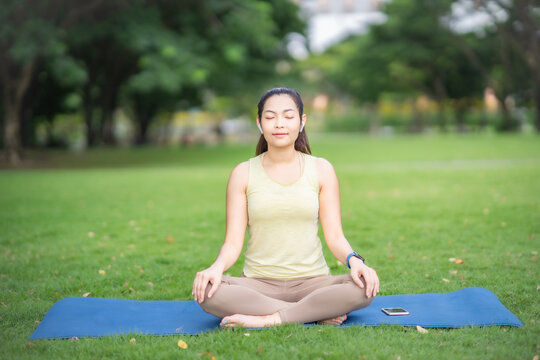 Young Asian Woman holding smartphone and ear bud sitting on yoga mat listening music do meditation with smile and happiness of paceful emotion at park
