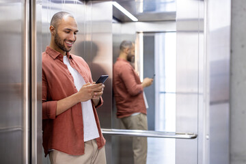 Man standing in elevator using smartphone, leaning against mirror wall, smiling effortlessly,...