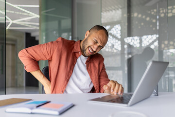 Man experiencing back pain while working at office desk with laptop. Concept of discomfort, posture issues, and need for ergonomic workspace during work. Focus on health.