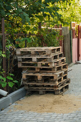 A tall stack of weathered wooden pallets rests beside a stone pathway, surrounded by vibrant greenery and shadows cast by nearby plants on a sunny day.