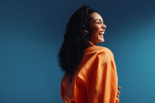 Young woman laughing while listening to a podcast, vibrant orange on blue background emphasizing joy and modernity