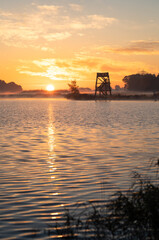 A wooden overlook tower at a lake during a tranquil, foggy sunrise in autumn.