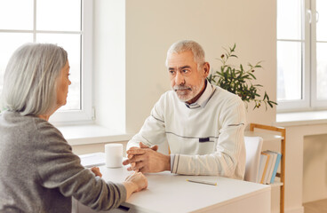 Obraz premium Elderly husband and wife, sitting together at a table at home, engage in a serious conversation. The senior couple talking about important matters, showing strong bond and communication as a couple.