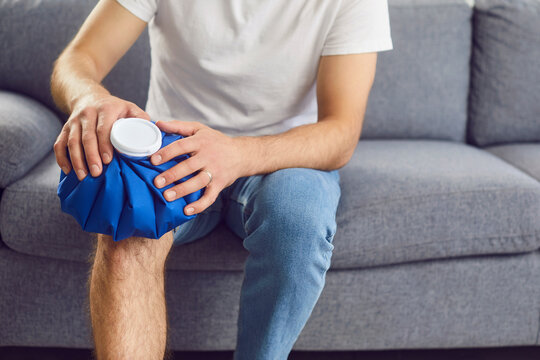 Unrecognizable man with a blue ice pack on his injured knee sitting on a gray couch at home. Concept of using a cold compress for pain and injury treatment