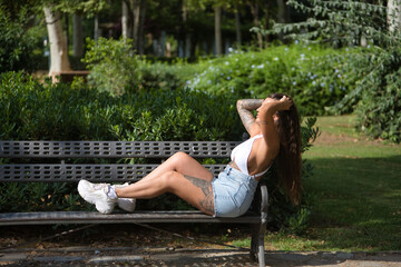 Young woman, brunette, beautiful, very tattooed, with white top and jeans, sitting on a park bench,...