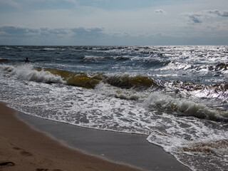 Seascape of the Baltic sea with big foamy waves in windy day with brown sand on the shore and blue sky above in sunlight