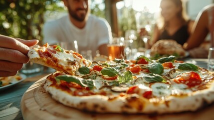 Friends enjoy a freshly baked pizza topped with basil and tomatoes on a wooden table in a sunlit outdoor space, capturing a sense of communal joy and culinary delight.