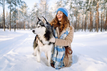A young woman in warm clothes walking with her husky dog in a picturesque snowy outdoor. Playing with pets. Friendship concept.