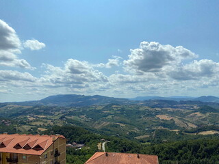 A panorama of a sunny blue sky decorated with patterns of illuminated snow-white clouds floating over the rooftops of San Marino to the mountain horizon.