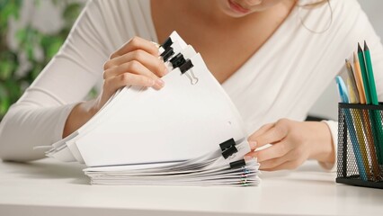 Woman sitting at office desk searching through overload stack of paper documents with binder and colorful paper clips. Bureaucracy business concept.