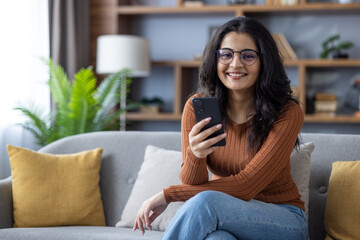 Portrait of a smiling young Indian woman in glasses sitting on the sofa at home, holding a mobile...