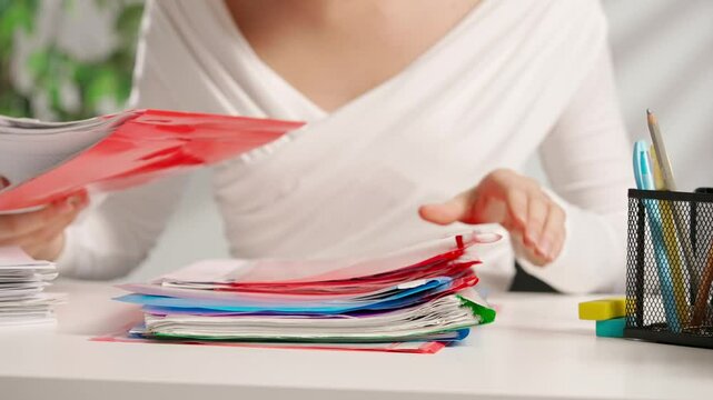 Woman sitting at office desk and searching through overload stack of colorful file folders full of paper documents sheets. Bureaucracy business concept.