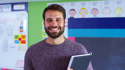 Portrait Of Male Elementary School Teacher Standing In Classroom Holding Folder