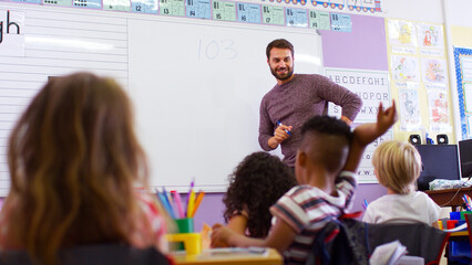 Male Teacher Standing At Whiteboard Teaching Maths Lesson To Elementary Pupils In School Classroom