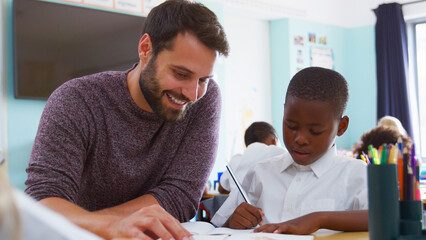 Male Elementary School Teacher Giving Pupil Wearing Uniform One To One Support In Classroom 
