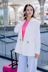 Vertical shot of serious business woman in a casual suit with a pink suitcase talking on a smartphone in modern train or bus station.