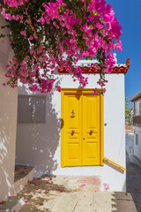 A striking yellow door stands out against the white walls, adorned with blooming bougainvillea, creating a picturesque setting in Hydra, Greece during the day.