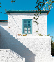 A charming traditional house in Hydra features striking white walls and a blue window, reflecting local architectural style under bright skies, surrounded by greenery.