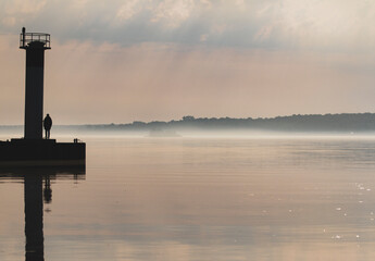 Fototapeta premium Silhouette of a lone man standing beside a small tower at the end of a pier in the early morning mist.