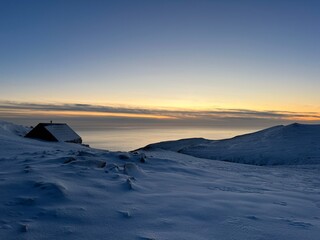 Seaside winter view. 
The photo captures a peaceful, snowy landscape at dusk. A small, dark cabin...