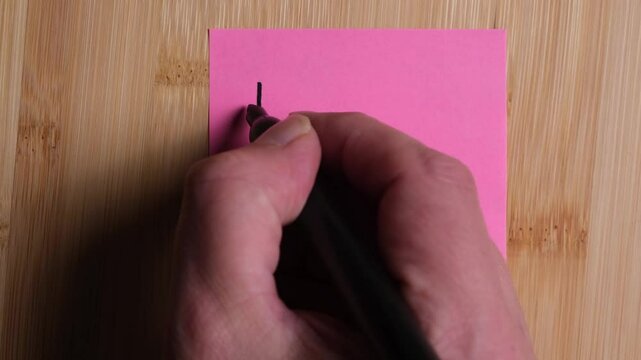 A person is writing on a sticky note on a wooden background. The person is writing I Love You, filmed in a slow motion style.