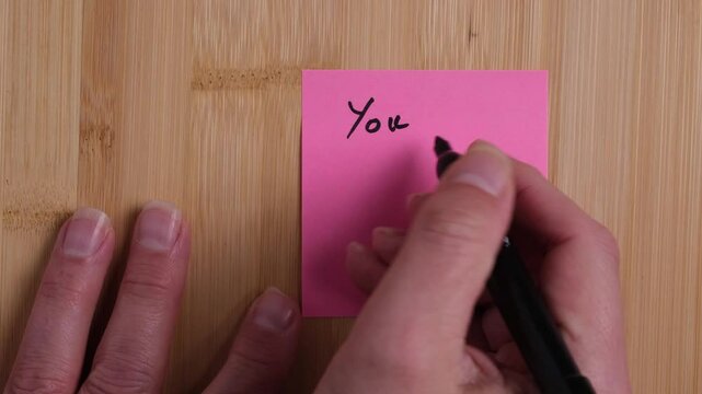 A person is writing on a sticky note on a wooden background. The person is writing You Are Wonderful. Concept, positivity, gratitude, affirmation.