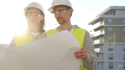 Architects or engineers wearing safety helmets and vests holding blueprint and discussing something...