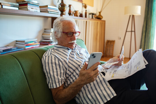 Elderly man reviewing bills and using smartphone on the couch