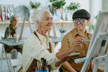 Senior Caucasian woman using brush and palette with aquarelle to paint on canvas while her mature...