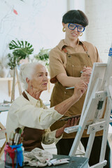 Vertical shot of young drawing teacher helping her senior silver haired woman to sketch on canvas...