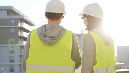 Two architects wearing safety hard hats and vests holding blueprint and discussing construction site project at sunrise, back view. Architecture, engineering and teamwork concept