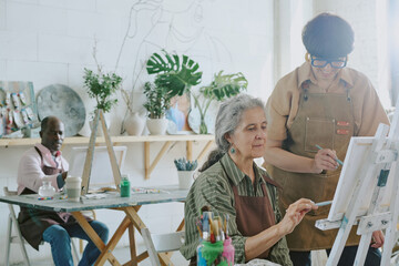 Young drawing teacher cooperating with her senior female student, they painting on canvas while...
