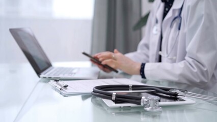 Stethoscope and clipboard are lying on the glass table while doctor woman is using a smartphone at the background. Medicine concept