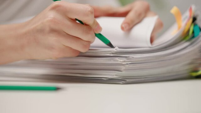 Woman sitting at office desk sorting and signing overload stack of paper documents with colorful clips with pencil, close up. Bureaucracy business concept.