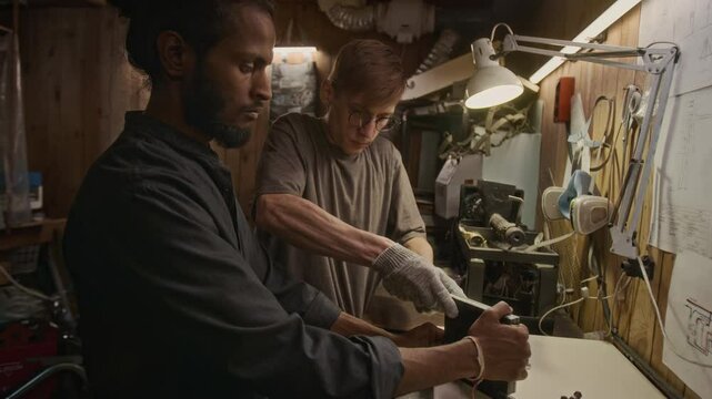 Medium shot of Caucasian mechanic disassembling metal component in warehouse, removing bolts with wrench, Indian male colleague helping, holding and turning it