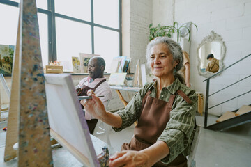 Medium close up of female pensioner doing strokes of paint brush during art session together with...