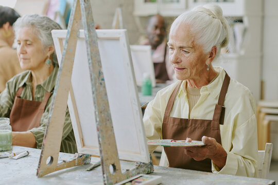 White Haired senior woman holding wooden palette with mixed paints while doing strokes on canvas